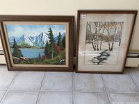 Overview photo of two framed paintings side by side, one oil painting of mountains and lake on the left, and one watercolor painting of snowy birch trees and creek on the right.