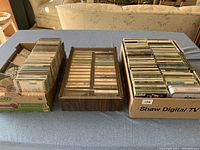 A wide view showing two cardboard boxes filled with CDs and a wooden cassette tray with 28 cassette tapes