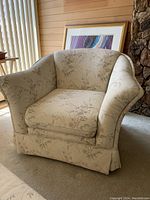 Front-side view of the beige Sklar Peppler armchair showing curved back, flared arms, floral upholstery, and skirted base.