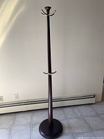 Full view of the tall wooden coat rack with brass hooks and circular base against a wall.