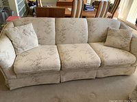 Full frontal view of the cream floral upholstered Sklar Peppler couch with three seat cushions and two throw pillows on a carpeted floor in a dining/living room.