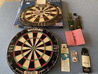 Overall lot showing new Winmau BDO dartboard, original box, assorted darts, darts case, rules booklet, razor sharpener, and spare tips.