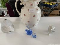 Set of items including a large floral jug, white bird vase, blue crystal swan, clear crystal swan, and white elephant figurine on a table.