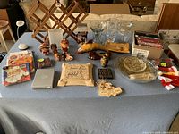Full overview of lot on table including dolls, serving ware, clear glass plates, figurines, decorative plaques, DVDs, and more.