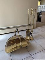 Brass fireplace tool set stand with brush, shovel, poker, and tongs, along with the brass wood log holder with black handle, arranged on the floor near a baseboard heater.