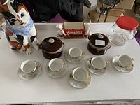 Overview of kitchen collectibles and ceramics including cat cookie jar, demitasse cup set, covered pots, Campbell's soup dish, and Kraft jar.