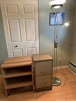 Photo showing wooden printer stand, metal filing cabinet, and floor lamp in corner of room near closet door and baseboard heater.