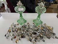 Wide view of two green glass oil lamps placed on a white surface in front, with a large group of collector spoons of various designs spread out in front of the lamps.