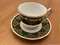 Bone china cup and saucer with dark green and gold ornamental pattern and gold rim on cup and saucer edges, displayed on wooden surface.