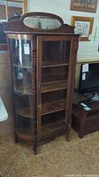 Full view of wooden curio cabinet showing front with oval mirror on top, door missing glass panel, curved glass side panel and shelves inside.
