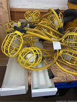 Extended string of yellow cage-protected construction lights with bulbs on wooden surface along with two fluorescent light fixtures in boxes.