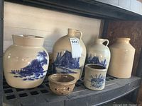 Wide shot showing all six earthenware pieces on metal shelf, including two large jugs with blue rural landscape paintings, a plain large crock, a small bowl, a small container, and a crock with blue flower basket design.