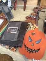 Photo showing a large orange wooden pumpkin wall hanging with cut-out eyes and jagged mouth, featuring a painted spider.
