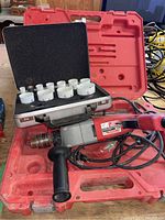 Photo of Milwaukee hammer drill in red case with black auxiliary handle and power cord, and open black case of white hole saw drill bits.