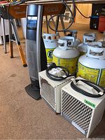 Overall view showing two white metal heaters and Bionaire upright tower fan beside several propane tanks in background.