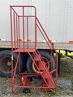 Red portable metal staircase with five perforated steps and safety cage, shown standing upright outdoors next to truck tires.