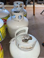 Two white 7.7 kg propane tanks with green and yellow labels and metal valve protective guards, photographed indoors on beige carpet.