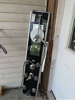 Full view of black Pulse shower panel with multiple chrome sprayers and controls leaning against wall.