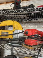 Photo showing the three tools on a wire shelf: Dewalt yellow orbital jig saw DW317, red Black & Decker sander, and grey Makita 5 inch sander B05030.
