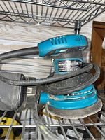 Makita BO5030 palm sander showing teal body, black grip, and attached power cord resting on metal shelving.