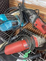 Three corded power tools including a blue Makita Skil saw, and two red Milwaukee tools, placed together on a metal rack, showing corded power connections and dust accumulation.