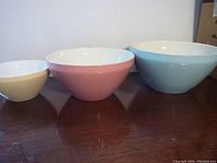 Three graduated pastel mixing bowls in cream, pink and blue, arranged from smallest to largest, on dark wooden surface.
