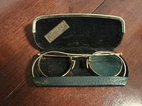 Antique gold-tone metal frame glasses with one green-tinted lens intact, placed inside a black textured case with silver metal corners on wooden table.