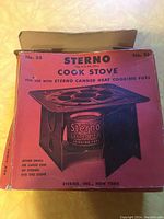 Red original box labeled 'Sterno Cook Stove' with text indicating use with Sterno canned heat fuel, showing some wear.