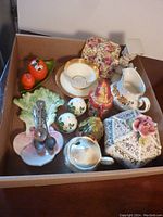Photo showing various ceramic and china items packed in a box including a decorative floral box, cup and saucer, pitchers, and ceramic figurines.
