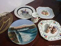 Overall lot showing 4 china plates, yellow glass relish dish, antique shaving mug, and two figural bottle stoppers arranged on wood surface.