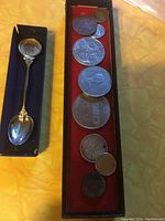 Photo showing the souvenir spoon in a display box with the collection of coins and medallions arranged on red felt.