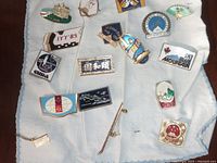 Photo showing 16 assorted metal enamel pins with foreign and ski themes arranged on white fabric.