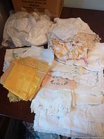 Box containing a pile of assorted fabric and linen pieces including doilies and crocheted items, mostly white and cream colored.