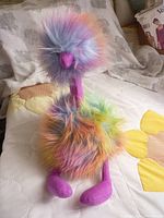 Full view of the Jellycat Rainbow Pompom Ostrich plush showing its pastel rainbow fluffy fur and purple feet and beak on a white quilted background.