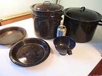 Set of black enamel kitchenware displayed with soda can for scale including two large pots with lids, steamer insert, two pie plates, and a funnel.