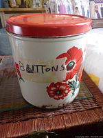 Large vintage tin with red lid and floral decoration showing the front of the tin with visible signs of rust and peeling tape labeled 'BUTTONS'.