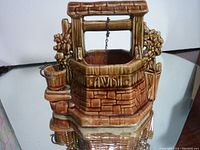 Front and side view of the brown glazed McCoy wishing well planter showing the brick texture, floral details, and attached small bucket on chain under crossbar handle.