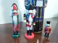 Three mini wooden nutcrackers in different uniform colors and a large nutcracker-shaped tin in the background.