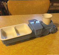 Front view of blue children's eating set shaped like a train with a cup in the smokestack section and a two-compartment tray in the cargo car.
