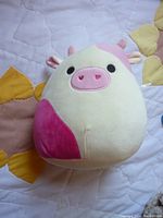 Front view of the pink and cream cow Squishmallow plush toy on quilted fabric.