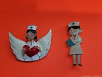 Photo showing two nurse enamel brooches, one with wings holding a heart, the other holding a clipboard with a white hat and red cross.