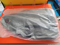 Black crossbody shoulder bag sealed in clear plastic packaging, shown on orange surface