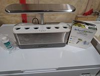 AeroGarden hydroponic growing system with attached LED light bar and eight planting holes. Also shown is the AeroGarden Gourmet Herbs seed kit box and a small bottle of liquid plant food next to the unit on a white surface.