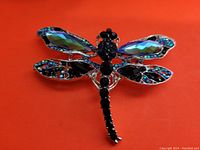 Close-up image of large dragonfly brooch with multicolored faceted crystals on wings and black glass stones forming the body, set on silver-tone metal against orange background.