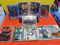 Photo showing all 10 F1 trading cards laid out on an orange surface with a yellow background. Cards include driver portraits of Lewis Hamilton, Max Verstappen, and others, as well as one card of a Mercedes race car on track.