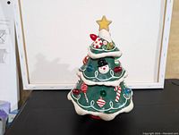 Green vintage ceramic Christmas tree with white borders, colorful candy cane and ornament decorations, snowman face, and yellow star top, in front on white floor and wall.