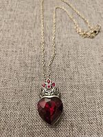 Close-up photo of vintage red amulet necklace showing heart-shaped red gem pendant with silver crown and three small red stones, on gold-toned chain.