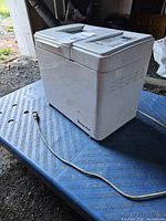 White Magic Chef bread maker on blue textured surface, showing side and rear with power cord.
