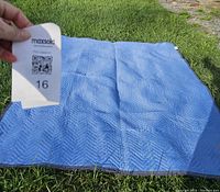 Blue quilted moving blanket lying flat on grass, showing texture and color, with a hand holding a MaxSold ticket marked 16 for lot identification.
