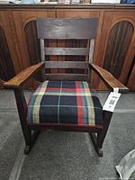 Front view of wooden rocking chair showing ladder back design and padded seat cushion with plaid pattern.